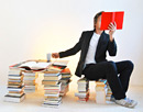 Link zur Seite „www.praktisch-unschlagbar.de“ (Ein junger Mann sitzt zwischen Bücherstapeln und liest ein rotes Buch)