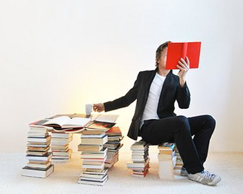 Ein junger Mann sitzt zwischen Bücherstapeln und liest ein rotes Buch