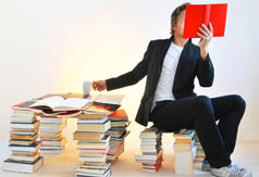 Link zur Seite „Lexikon“ (Ein junger Mann sitzt auf einem Bücher-Stapel und liest ein Buch)