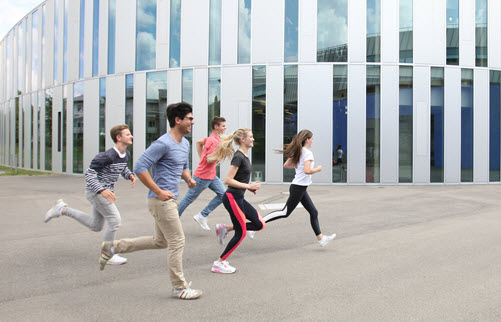 Zwei Schülerinnen und drei Schüler laufen an einer modernen Gebäudefassade aus Glas und Metall vorbei