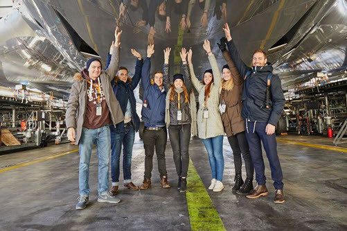 Die Mitglieder der Schülerfirma Promising Six stehen in einer Lufthansa Werkshalle in Frankfurt am Main unter dem Rumpf einer Flugzeugs und strecken die Hände hoch
