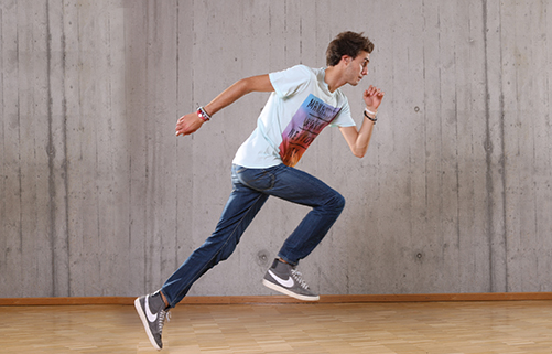 Junge in Jeans, Turnschuhen und T-Shirt vor einer grauen Wand läuft auf Holzparkett