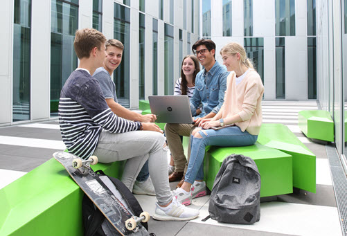Gruppe Schülerinnen und Schüler mit Laptop sitzen auf grünen Sitzgelegenheiten in einem Gebäudehof