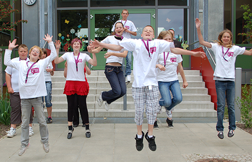 Schüler mit GRÜNDERKIDS T-Shirt vor einer Schule springen in die Luft