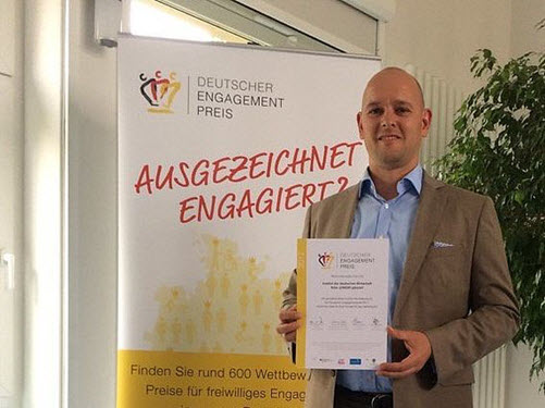 IW JUNIOR Leiter Felix Hettig steht vor dem Plakat des Deutschen Engagementpreises und hält die Urkunde in seinen Händen