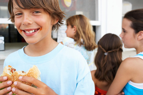 Kleiner Junge lächelt fröhlich in die Kamera und hält einen angebissenen Hamburger in der Hand. 