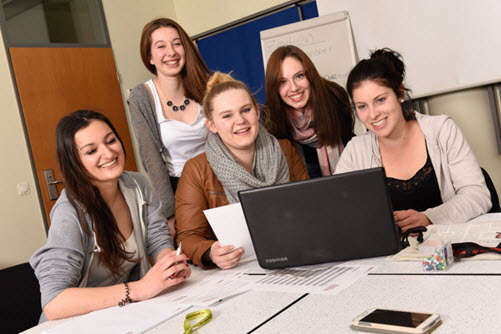Fünf Schülerinnen schauen gemeinsam auf einen Laptop, der auf dem Tisch in einem Klassenraum steht