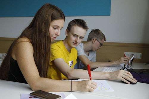 Eine Schülerin und zwei Schüler sitzen mit Laptop an einem Schreibtisch im Klassenraum und arbeiten