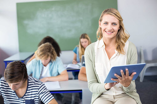 Lehrerin mit Tablet in den Händen sitzt auf einem Tisch vor Ihren Schülern in einem Klassenraum